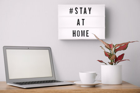 Laptop, cup and lightbox with hashtag STAY AT HOME indoors. Message to promote self-isolation during...