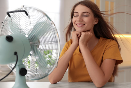 Woman enjoying air flow from fan at table in kitchen. summer heatの写真素材