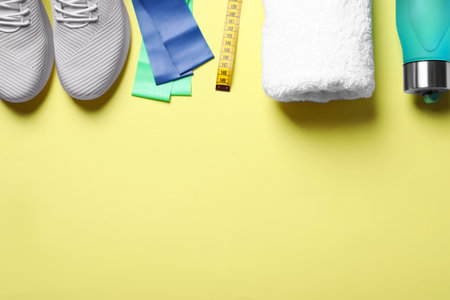 Sneakers, fitness elastic band, measuring tape, towel and bottle of water on yellow background, flat lay. Space for textの写真素材