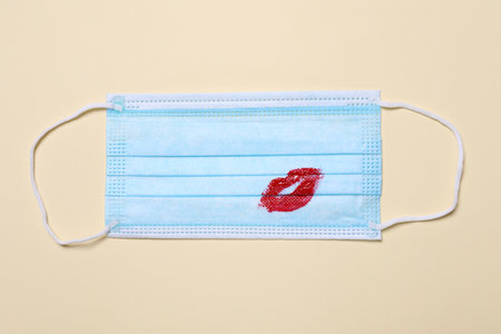 Medical mask with lipstick mark on beige background, top view