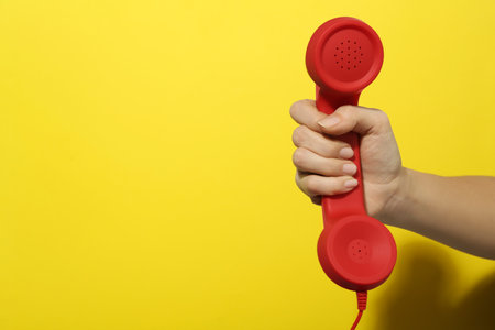 Closeup view of woman holding red corded telephone handset on yellow background, space for text. hotline conceptの写真素材