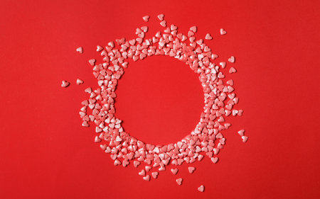 Round frame made of bright heart shaped sprinkles on red background, flat lay. Space for textの写真素材