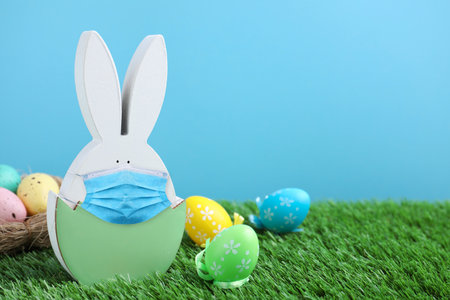 COVID-19 pandemic. Easter bunny figure in protective mask and dyed eggs on green grass against light...
