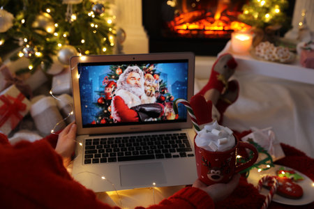 MYKOLAIV, UKRAINE - DECEMBER 23, 2020: Woman with sweet drink watching The Christmas Chronicles movie on laptop near fireplace at home, closeup. Cozy winter holidays atmosphereのeditorial素材