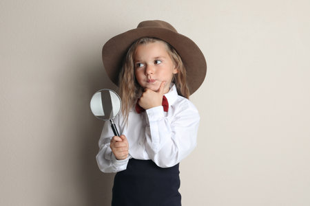 Cute little child in hat with magnifying glass playing detective on beige backgroundの写真素材