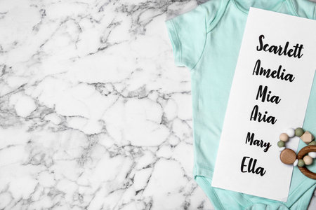 Bodysuit with list of baby names and toy on white marble background, top view. Space for text