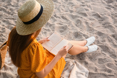 Young woman reading book on sandy beach, back viewの写真素材