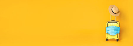 COVID-19 pandemic, travel during coronavirus outbreak. Suitcase with protective mask and hat on yellow background, space for textの写真素材