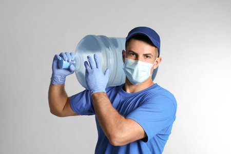Courier in face mask with bottle of cooler water on gray background.の写真素材