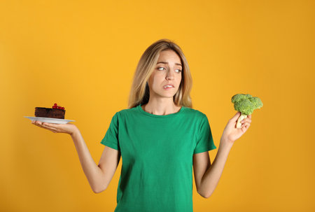 Woman choosing between cake and healthy broccoli on yellow backgroundの写真素材