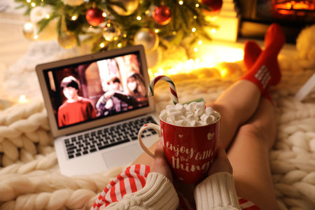 MYKOLAIV, UKRAINE - DECEMBER 23, 2020: Woman with sweet drink watching Harry Potter and Philosopher's Stone movie on laptop near fireplace at home, closeup. Cozy winter holidays atmosphereのeditorial素材
