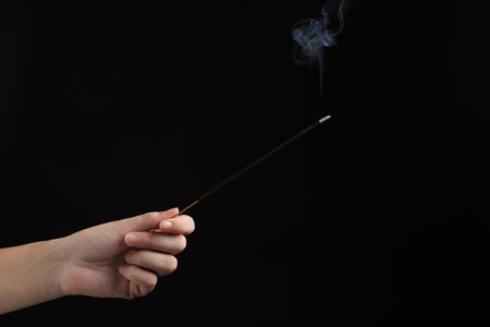 Woman holding smoldering incense stick on black background, closeup. Space for textの写真素材