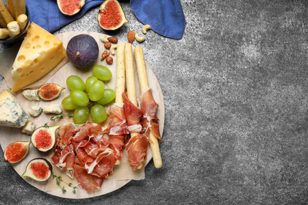 Board with delicious figs, cheese, grapes and bread with proscuitto on gray table, flat lay. Space for textの写真素材