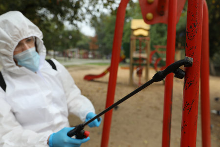 Woman wearing chemical protective suit at playground, focus on disinfectant sprayer. Preventive measure during coronavirus pandemicの写真素材