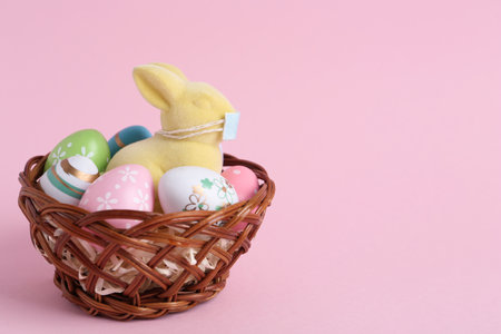 Cute bunny figure with protective mask and eggs in wicker basket on pink background, space for text. Easter holiday during COVID-19 quarantineの写真素材