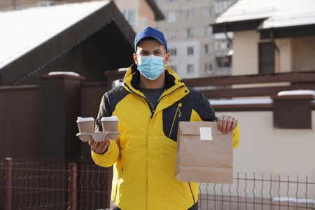 Courier in medical mask holding takeaway food and drinks near house outdoors. Delivery service...