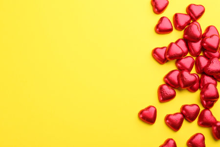 Heart shaped chocolate candies in red foil on yellow background, flat lay. Space for textの写真素材