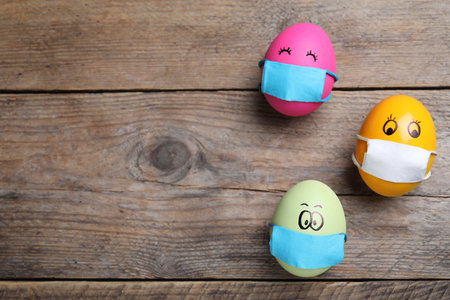 Bright eggs in protective masks on wooden table, flat lay with space for text. Easter holiday during   quarantineの写真素材