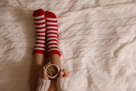 Woman in warm socks with cup of hot drink relaxing on knitted blanket, top view. Space for textの写真素材