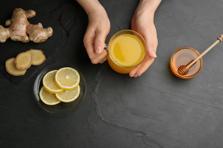 Woman holding cup of immunity boosting drink at dark table with ingredients, top view. Space for textの写真素材