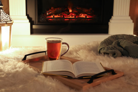 Cup of tea and book on fuzzy rug near fireplace at home. Cozy atmosphereの写真素材