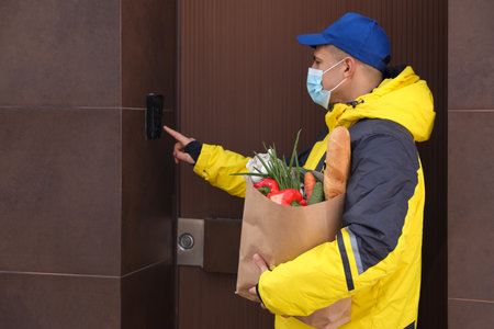 Courier in medical mask holding paper bag with groceries and ringing doorbell outdoors. Delivery service during quarantine due to Covid-19 outbreakの写真素材