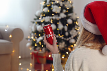 MYKOLAIV, UKRAINE - January 01, 2021: Woman in Santa hat with can of Coca-Cola against blurred Christmas tree at home, closeupのeditorial素材