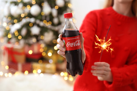 MYKOLAIV, UKRAINE - January 01, 2021: Woman with bottle of Coca-Cola and sparkler against blurred Christmas tree, closeupのeditorial素材