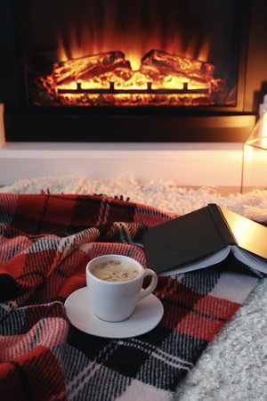 Cup of hot drink and book on blanket near fireplace indoors. magic atmosphereの写真素材