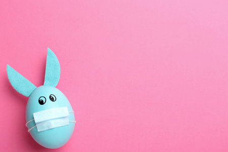Painted egg decorated with bunny ears, protective mask and space for text on pink background, top view. Easter holiday during COVID-19 quarantineの写真素材