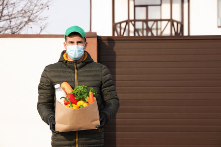 Courier in medical mask holding paper bag with groceries near house outdoors. Delivery service during quarantine due to Covid-19 outbreakの写真素材