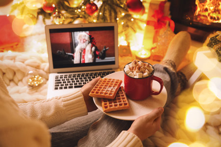 MYKOLAIV, UKRAINE - DECEMBER 23, 2020: Woman with cocoa and wafers watching The Christmas Chronicles movie on laptop near fireplace at home, closeup. Cozy winter holidays atmosphereのeditorial素材