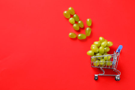 Arrow made of grapes pointing at small shopping cart with berries on red background, flat lay. Space for textの写真素材