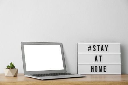 Laptop and lightbox with hashtag STAY AT HOME on wooden table. Message to promote self-isolation during pandemicの写真素材
