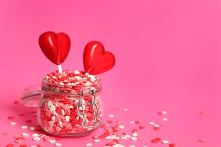 Sweet heart shaped lollipops and sprinkles in glass jar on pink background, space for text. Valentine's day celebrationの写真素材