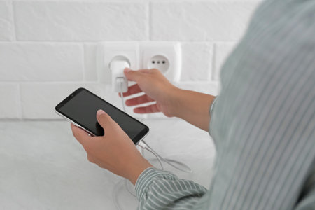 Woman with mobile phone plugging charger in socket on white brick wall, closeup