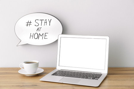 Laptop, cup of coffee and speech bubble with hashtag STAY AT HOME on white wall.の写真素材