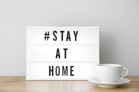 Cup and light box with hashtag STAY AT HOME on wooden table near white wall.の写真素材