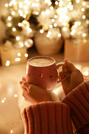 Woman with cup of drink and blurred Christmas lights on background, closeupの写真素材