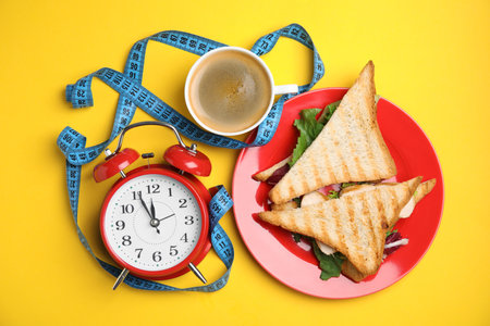 Flat lay composition with tasty sandwiches and alarm clock on yellow background. Nutrition regimenの写真素材