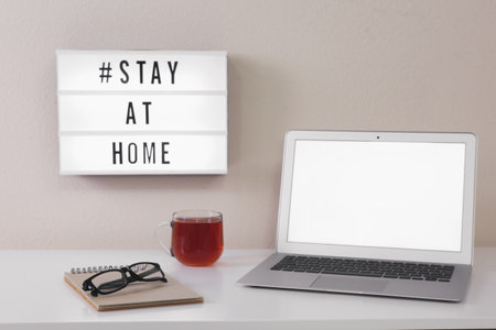 Laptop, cup of tea and lightbox with hashtag STAY AT HOME on white wall. Message to promote self-isolation during COVIDâ19の写真素材