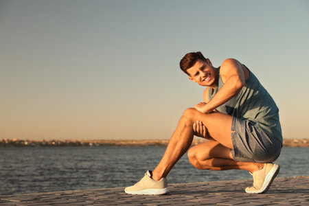 Man in sportswear having knee problems near river at sunsetの写真素材