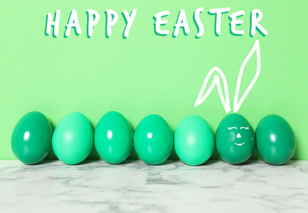One egg with drawn face and ears as Easter bunny among others on white marble table against green backgroundの写真素材