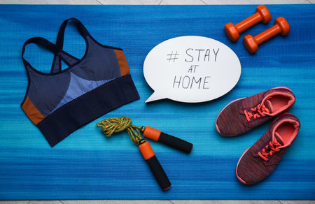 Sport equipment and speech bubble with hashtag STAY AT HOME on blue yoga mat, flat lay.