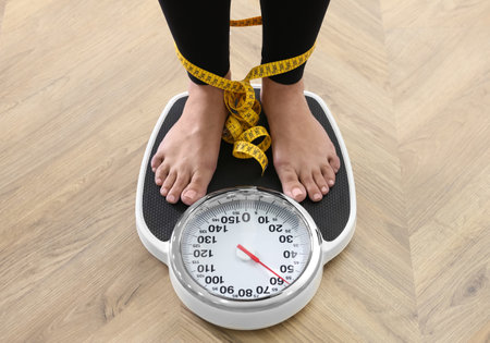 Woman tied with measuring tape using scale on floor, closeup. Overweight problem after New Year partyの写真素材