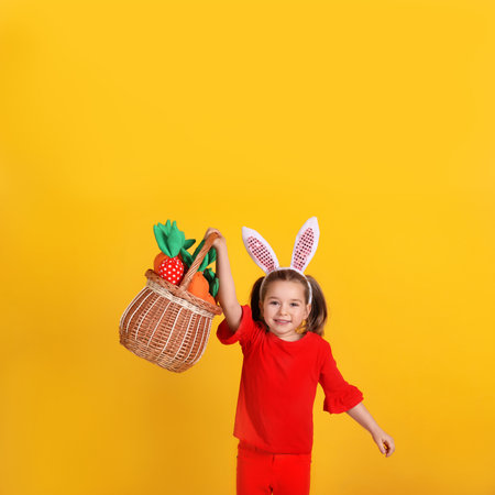 Happy little girl with bunny ears holding wicker basket full of toy carrots on orange background. easter celebrationの写真素材