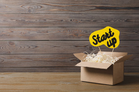 Open cardboard box and sign with words Start Up on wooden table, space for text