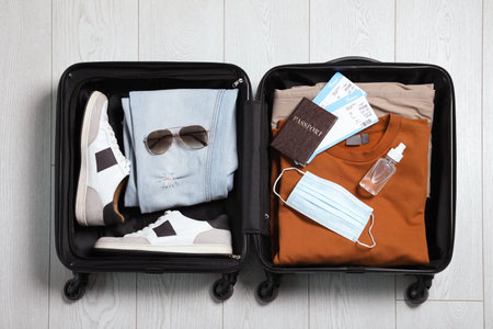 Open suitcase with passport, tickets, sanitizer and protective mask on wooden floor, top view. Travel during quarantineの写真素材