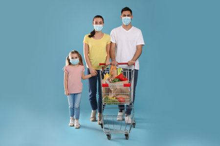 Family with protective masks and shopping cart full of groceries on light blue backgroundの写真素材