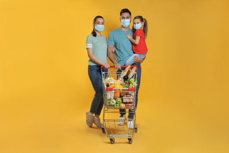 Family with protective masks and shopping cart full of groceries on yellow backgroundの写真素材